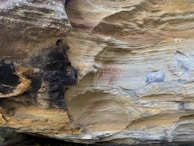A close-up view of rocky surfaces with a combination of dark and light tones. There are visible layers and textures that suggest erosion and weathering. Red markings, possibly ancient or symbolic, appear faintly on the rock's lighter surface.
