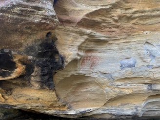 A close-up view of rocky surfaces with a combination of dark and light tones. There are visible layers and textures that suggest erosion and weathering. Red markings, possibly ancient or symbolic, appear faintly on the rock's lighter surface.