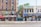 A street view of a city block featuring various local businesses. On the left, there is a brick building with signs advertising Medicare services, featuring blue awnings. To the right, a two-story building houses a beauty and hair salon. On the ground floor, a laundromat and a local market with signs advertising various goods can be seen. Pedestrians are walking along the sidewalk, and a tree is planted in the pavement. An empty street runs parallel to the buildings.
