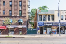 A street view of a city block featuring various local businesses. On the left, there is a brick building with signs advertising Medicare services, featuring blue awnings. To the right, a two-story building houses a beauty and hair salon. On the ground floor, a laundromat and a local market with signs advertising various goods can be seen. Pedestrians are walking along the sidewalk, and a tree is planted in the pavement. An empty street runs parallel to the buildings.