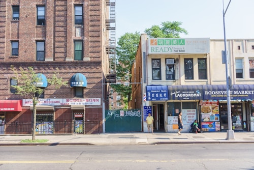 A street view of a city block featuring various local businesses. On the left, there is a brick building with signs advertising Medicare services, featuring blue awnings. To the right, a two-story building houses a beauty and hair salon. On the ground floor, a laundromat and a local market with signs advertising various goods can be seen. Pedestrians are walking along the sidewalk, and a tree is planted in the pavement. An empty street runs parallel to the buildings.