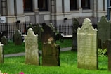 Several old headstones stand on a lush green lawn, with a wrought iron fence and a stone building visible in the background. The tombstones are weathered, with inscriptions partially faded, indicating historical significance.
