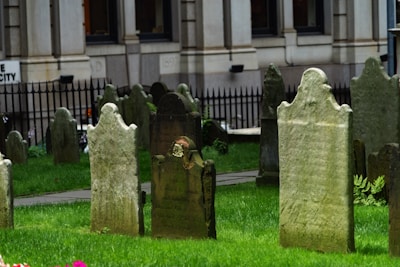 Several old headstones stand on a lush green lawn, with a wrought iron fence and a stone building visible in the background. The tombstones are weathered, with inscriptions partially faded, indicating historical significance.