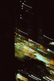 A nighttime cityscape with blurred lights from moving vehicles and illuminated buildings. The image appears to capture motion, creating streaks of light across the frame. Dark areas dominate the image, providing strong contrast with the vibrant light trails from the cars.