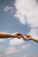 Two hands reaching towards each other against a backdrop of a clear blue sky with scattered clouds, conveying a sense of connection and intimacy.