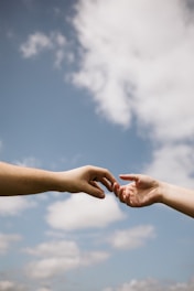 Two hands reaching towards each other against a backdrop of a clear blue sky with scattered clouds, conveying a sense of connection and intimacy.