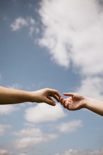Two hands reaching towards each other against a backdrop of a clear blue sky with scattered clouds, conveying a sense of connection and intimacy.