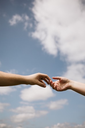 Two hands reaching towards each other against a backdrop of a clear blue sky with scattered clouds, conveying a sense of connection and intimacy.