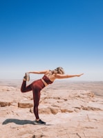 A person wearing maroon fitness clothing is performing a yoga or stretching pose on a rocky plateau. The person is balancing on one leg while holding the other leg behind them, with one arm extended forwards. The background shows an expansive, bright blue sky and a vast, barren landscape.