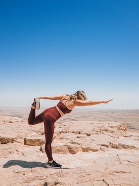 A person wearing maroon fitness clothing is performing a yoga or stretching pose on a rocky plateau. The person is balancing on one leg while holding the other leg behind them, with one arm extended forwards. The background shows an expansive, bright blue sky and a vast, barren landscape.