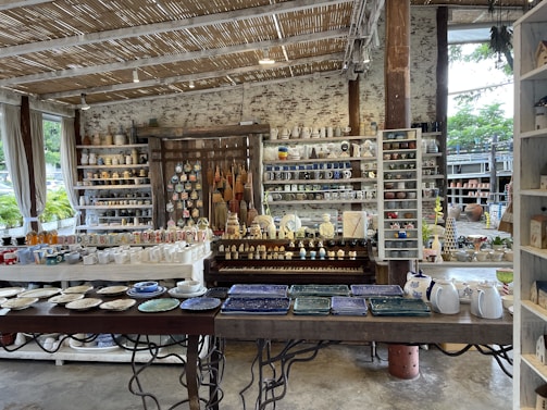 A rustic indoor shop displays a variety of ceramic and pottery items, including mugs, plates, bowls, and decorative figures. The shelves are filled with neatly arranged, colorful pottery items. The shop features a natural aesthetic with wooden shelves, a thatched-style ceiling, and large windows letting in natural light. Plants can be seen outside through the windows.