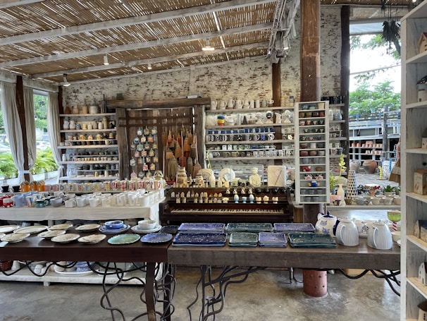 A rustic indoor shop displays a variety of ceramic and pottery items, including mugs, plates, bowls, and decorative figures. The shelves are filled with neatly arranged, colorful pottery items. The shop features a natural aesthetic with wooden shelves, a thatched-style ceiling, and large windows letting in natural light. Plants can be seen outside through the windows.