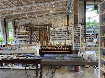 A rustic indoor shop displays a variety of ceramic and pottery items, including mugs, plates, bowls, and decorative figures. The shelves are filled with neatly arranged, colorful pottery items. The shop features a natural aesthetic with wooden shelves, a thatched-style ceiling, and large windows letting in natural light. Plants can be seen outside through the windows.