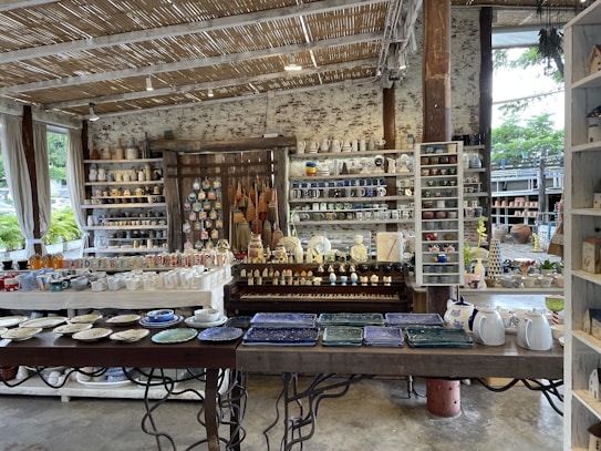 A rustic indoor shop displays a variety of ceramic and pottery items, including mugs, plates, bowls, and decorative figures. The shelves are filled with neatly arranged, colorful pottery items. The shop features a natural aesthetic with wooden shelves, a thatched-style ceiling, and large windows letting in natural light. Plants can be seen outside through the windows.
