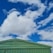 A corrugated metal roof of a green building is positioned against a vibrant sky filled with fluffy white clouds. The structure has a geometric design with sharp lines and angles, providing a contrast to the organic and soft shapes of the clouds above.