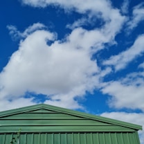 A corrugated metal roof of a green building is positioned against a vibrant sky filled with fluffy white clouds. The structure has a geometric design with sharp lines and angles, providing a contrast to the organic and soft shapes of the clouds above.