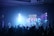 A concert or worship setting with a band performing on stage in front of an audience. The stage features bright lights and a large screen displaying the word 'JESUS' along with lyrics in both English and another language. The crowd is engaged, with some raising their hands.