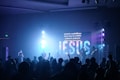 A concert or worship setting with a band performing on stage in front of an audience. The stage features bright lights and a large screen displaying the word 'JESUS' along with lyrics in both English and another language. The crowd is engaged, with some raising their hands.