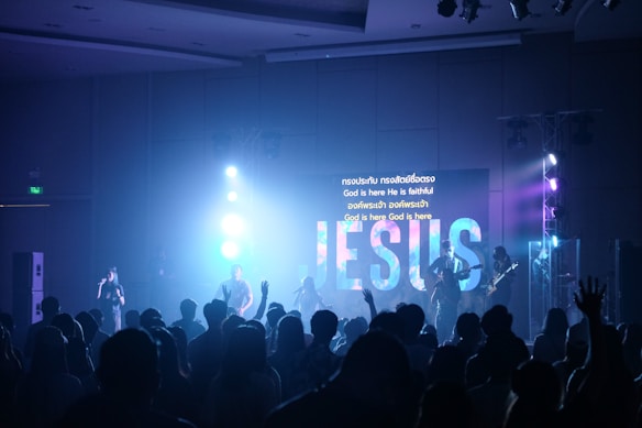 A concert or worship setting with a band performing on stage in front of an audience. The stage features bright lights and a large screen displaying the word 'JESUS' along with lyrics in both English and another language. The crowd is engaged, with some raising their hands.