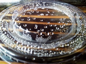 A close-up view of a transparent plastic lid with water droplets scattered on its surface. The background shows a wooden surface with distinct lines visible through the lid, emphasizing the clarity of the plastic and the droplets.
