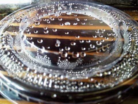 A close-up view of a transparent plastic lid with water droplets scattered on its surface. The background shows a wooden surface with distinct lines visible through the lid, emphasizing the clarity of the plastic and the droplets.