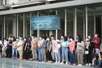 A group of people stands closely together by the edge of a pool, all wearing face masks. They appear to be attending an event related to baptism, as indicated by the banner above their heads. The individuals are casually dressed and are facing towards the water, suggesting anticipation or participation in the ceremony.