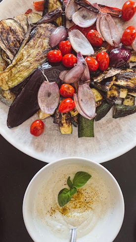 A plate filled with a variety of grilled vegetables like eggplant, cherry tomatoes, and onion slices. Below it, a bowl contains a creamy white dip garnished with basil leaves and a sprinkle of spices on top.