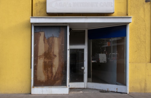 A closed storefront with yellow walls and a weathered sign on top. One window is boarded up with brown paper, and the other is a glass door with reflections of an empty street and a van. The entrance appears neglected and out of use.