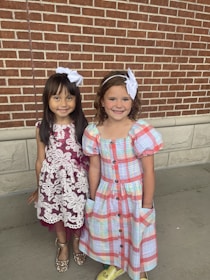 Two young girls stand together in front of a brick wall. Both girls have white bows in their hair. The girl on the left is wearing a maroon dress with white lace detailing and sparkly gold shoes. The girl on the right is wearing a colorful plaid dress with puffy sleeves and yellow shoes. They are both smiling and appear to be happy.