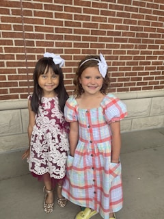 Two young girls stand together in front of a brick wall. Both girls have white bows in their hair. The girl on the left is wearing a maroon dress with white lace detailing and sparkly gold shoes. The girl on the right is wearing a colorful plaid dress with puffy sleeves and yellow shoes. They are both smiling and appear to be happy.