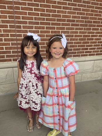 Two young girls stand together in front of a brick wall. Both girls have white bows in their hair. The girl on the left is wearing a maroon dress with white lace detailing and sparkly gold shoes. The girl on the right is wearing a colorful plaid dress with puffy sleeves and yellow shoes. They are both smiling and appear to be happy.