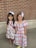 Two young girls stand together in front of a brick wall. Both girls have white bows in their hair. The girl on the left is wearing a maroon dress with white lace detailing and sparkly gold shoes. The girl on the right is wearing a colorful plaid dress with puffy sleeves and yellow shoes. They are both smiling and appear to be happy.