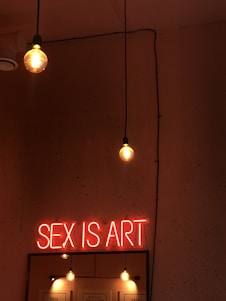 A dimly lit room with hanging light bulbs and a neon sign displaying the phrase 'SEX IS ART'. The background is a textured wall, and the lighting gives a warm, intimate atmosphere.