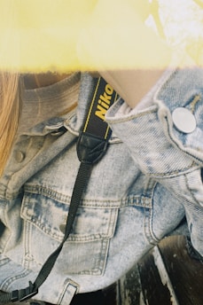 A close-up view of a person wearing a light denim jacket. A camera strap with the word 'Nikon' is partially visible, draped across the shoulder. The image has a yellowish tint at the top, likely due to lighting effects or a filter.