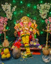 A colorful idol of the Hindu deity Ganesha adorned with garlands of yellow flowers, sitting on a decorated pedestal. The background features a lush green foliage with illuminated elements and white and pink floral arrangements. Two brass oil lamps are placed on either side of the idol. Fruits, coconuts, and other offerings are arranged in front of the statue.