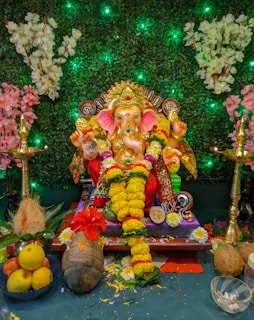 A colorful idol of the Hindu deity Ganesha adorned with garlands of yellow flowers, sitting on a decorated pedestal. The background features a lush green foliage with illuminated elements and white and pink floral arrangements. Two brass oil lamps are placed on either side of the idol. Fruits, coconuts, and other offerings are arranged in front of the statue.