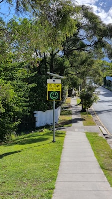 A sunny street scene with a sidewalk, bordered by grass and trees. A sign with a smiley face and the text 