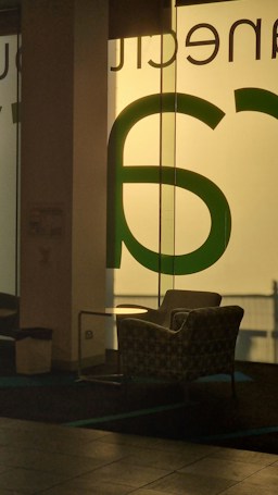 A cozy armchair sits in front of a large glass window with bold green lettering that allows soft light to filter through, creating a serene atmosphere. The space also includes a small side table next to the chair, and there's a waste bin nearby.