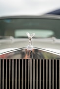 A close-up of a classic car's front emblem and grille, featuring a silver figurine above the car's badge. The emblem is polished and shiny, with intricate details visible. The focus is on the emblem while the background is softly blurred, suggesting luxury and elegance.