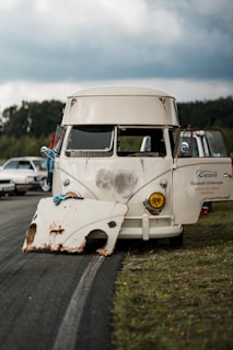 A vintage van is parked on the side of a road with its doors open. A detached, rusted vehicle part rests in front of it on the ground. The sky is overcast, and there are other cars and trees visible in the background.