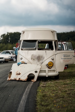 A vintage van is parked on the side of a road with its doors open. A detached, rusted vehicle part rests in front of it on the ground. The sky is overcast, and there are other cars and trees visible in the background.