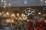 A variety of chandeliers and lamps are displayed in a shop window. The lighting fixtures range from traditional to modern designs, with some emitting a warm glow. Reflections of the street and nearby buildings are visible on the glass. In the foreground, there are red flowers and green foliage partially obscuring the view.