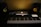 A music production setup featuring a MIDI keyboard controller in black and white keys, an illuminated laptop with an Apple logo, and two studio monitor speakers with yellow cones on a dark desk.