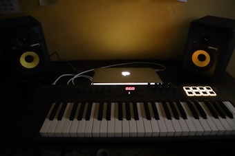 A music production setup featuring a MIDI keyboard controller in black and white keys, an illuminated laptop with an Apple logo, and two studio monitor speakers with yellow cones on a dark desk.