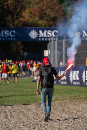 A person with a backpack and red cap walks on a gravel path while holding a lit flare emitting colorful smoke. In the background, there are more people gathered near a fence with a banner that reads 'MSC Cruises'. Trees surround the area.