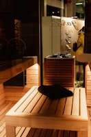 A wooden sauna interior with warm, ambient lighting. A black towel is placed on a wooden bench, and a wooden shelf holds rocks, likely for a heater. Outside, a person in a yellow coat can be seen through the glass, next to a wall with hanging objects.