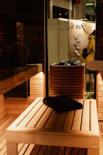 A wooden sauna interior with warm, ambient lighting. A black towel is placed on a wooden bench, and a wooden shelf holds rocks, likely for a heater. Outside, a person in a yellow coat can be seen through the glass, next to a wall with hanging objects.