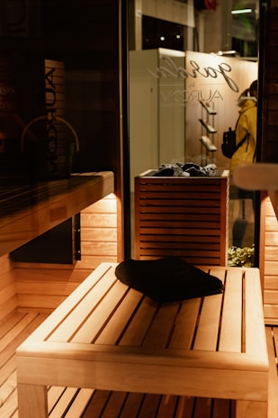 A wooden sauna interior with warm, ambient lighting. A black towel is placed on a wooden bench, and a wooden shelf holds rocks, likely for a heater. Outside, a person in a yellow coat can be seen through the glass, next to a wall with hanging objects.