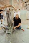 A man with a beard is kneeling on the ground next to a partially assembled metallic structure, possibly a shelf or display rack, inside a spacious industrial or workshop setting. He appears to be working on the assembly with tools nearby, including pliers. The environment is spacious with high ceilings and visible beams.