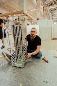 A man with a beard is kneeling on the ground next to a partially assembled metallic structure, possibly a shelf or display rack, inside a spacious industrial or workshop setting. He appears to be working on the assembly with tools nearby, including pliers. The environment is spacious with high ceilings and visible beams.
